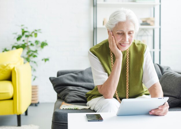 Lächelnde ältere Frau, die im Wohnzimmer betrachtet digitale Tablette sitzt
