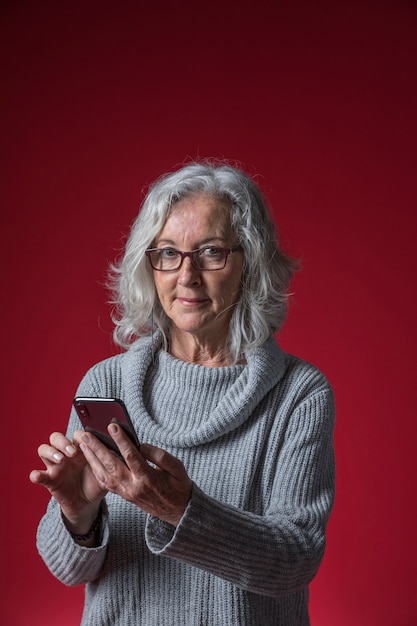 Kostenloses Foto lächelnde ältere frau, die in der hand das intelligente telefon schaut zur kamera gegen farbigen hintergrund hält