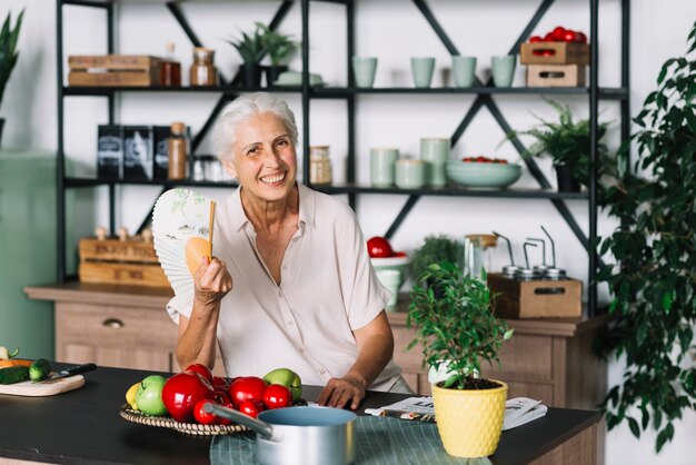 Lächelnde ältere Frau, die in der Hand den Fan sitzt hinter dem Küche worktop hält