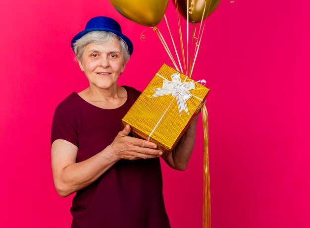 Lächelnde ältere Frau, die Partyhut trägt, hält Heliumballons und Geschenkbox lokalisiert auf rosa Wand
