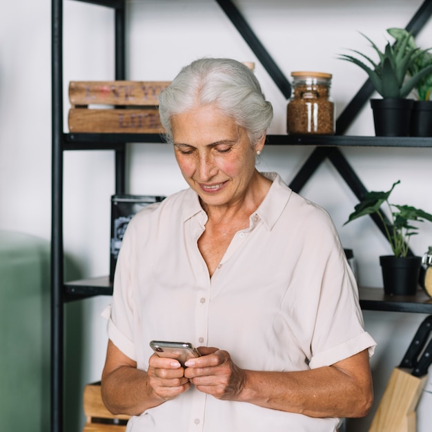 Lächelnde ältere Frau, die vor dem Regal unter Verwendung des Mobiles steht