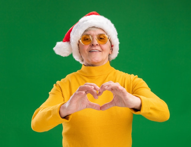 Lächelnde ältere Frau in Sonnenbrille mit Weihnachtsmütze gestikuliert Herzzeichen lokalisiert auf grünem Hintergrund mit Kopienraum