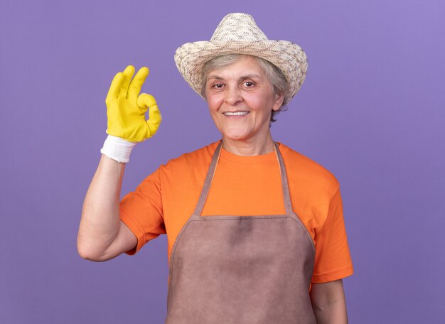 Lächelnde ältere Gärtnerin mit Gartenhut und Handschuhen gestikuliert ok singen isoliert auf lila Wand mit Kopierraum