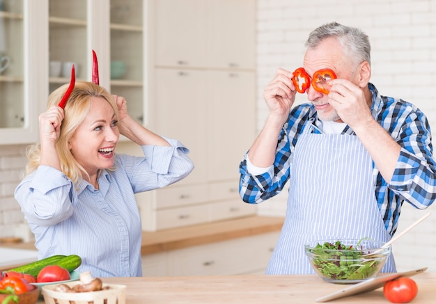 Lächelnde ältere Paare, die Spaß mit den Pfeffern und dem grünen Pfeffer des roten Paprikas stehen in der Küche machen