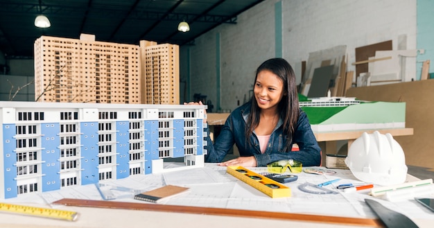 Lächelnde Afroamerikanerfrau nahe Modell des Gebäudes auf Tabelle mit Ausrüstungen