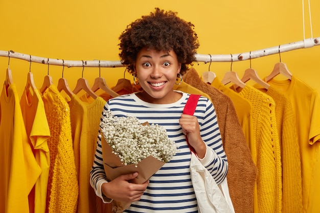 Lächelnde afroamerikanische Frau posiert gegen Schrank, wählt passendes neues Outfit, mag gelbe Farbe, trägt Tasche, hält Blumen, lächelt breit