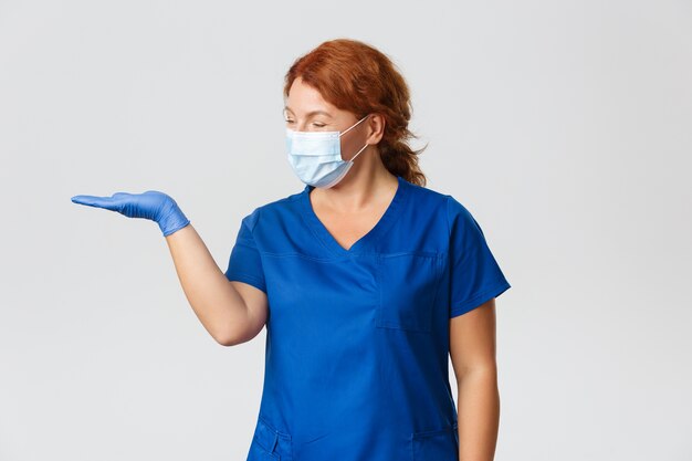 Lächelnde angenehme Ärztin, Krankenschwester, die glücklich aussieht, als würde sie etwas auf der Hand halten, Gesichtsmaske und Gummihandschuhe tragen