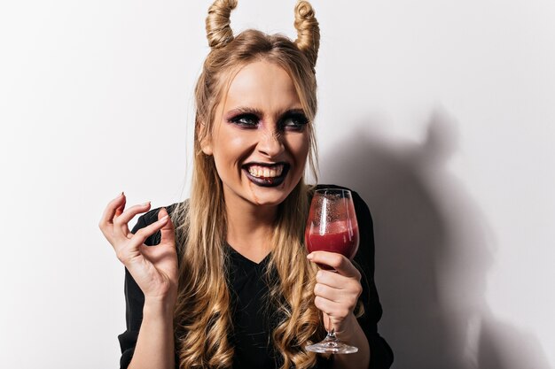 Lächelnde ansprechende Dame im Vampirkostüm, die Party genießt. Foto des lachenden Mädchens mit Kunstblut im Weinglas.