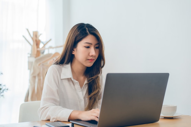 Lächelnde Asiatin der schönen Junge, die an Laptop während zu Hause im Büroarbeitsplatz arbeitet