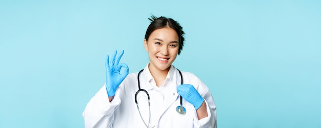 Lächelnde asiatische arztärztin, die das stethoskop berührt und zeigt, dass es okay ist, melden sie sich auf blauem hintergrund an