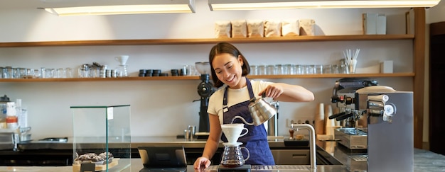 Lächelnde asiatische Barista schüttet heißes Wasser aus dem Kessel und braut Filterkaffee, der dahinter steht