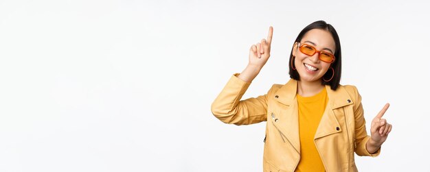 Lächelnde asiatische brünette Frau mit Sonnenbrille, die mit dem Finger seitlich nach links und rechts zeigt und Varianten zeigt, die mit Sonnenbrille auf weißem Hintergrund lachen und tanzen