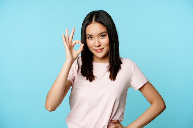 Lächelnde asiatische Frau, die ein Okay-Zeichen zeigt, gibt Zustimmung, empfiehlt etw guten Stand auf blauem Hintergrund