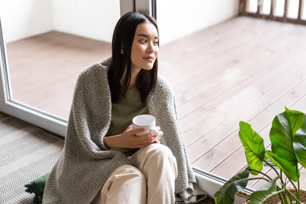 Lächelnde asiatische Frau, die mit verträumtem Gesicht aus dem Fenster schaut, morgens Kaffee trinkt und in der Nähe sitzt...