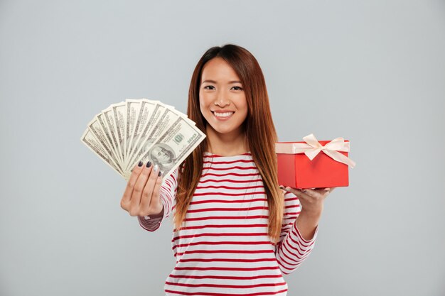 Lächelnde asiatische Frau im Pullover, der Geld und Geschenk über grauem Hintergrund hält