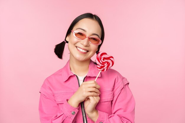 Lächelnde asiatische Frau mit Sonnenbrille, die Lolipop-Bonbons hält und Süßigkeiten isst und glücklich über rosa Hintergrund steht