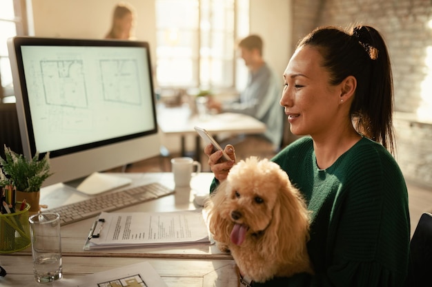 Lächelnde asiatische Geschäftsfrau mit einem Hund SMS auf dem Handy im Büro