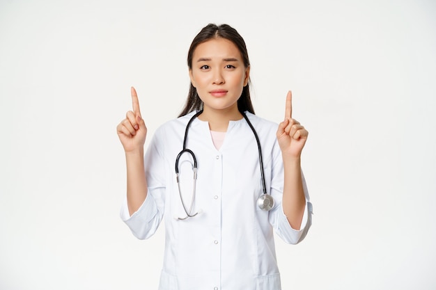Lächelnde asiatische Ärztin, die mit den Fingern nach oben zeigt und in die Kamera schaut, Informationen zur medizinischen Versorgung bereitstellt, weißer Hintergrund