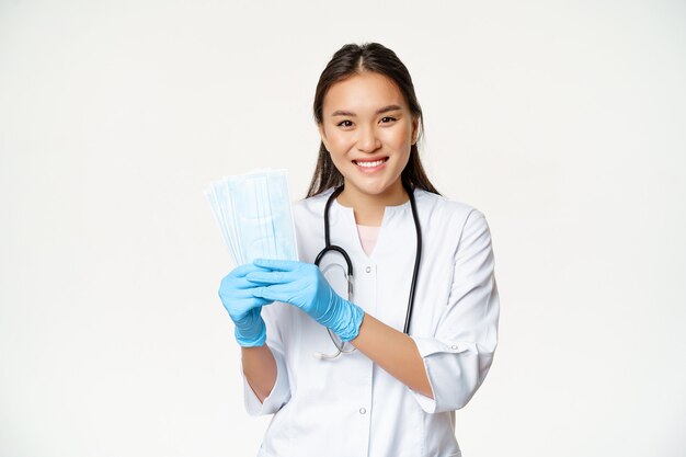 Lächelnde asiatische Ärztin, Krankenschwester, die medizinische Gesichtsmasken zeigt, sterile Gummihandschuhe trägt und auf weißem Hintergrund steht