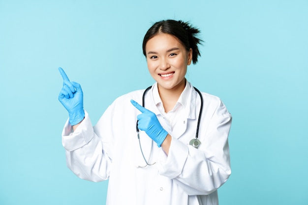 Lächelnde asiatische Ärztin, Therapeutin, die mit den Fingern auf die obere linke Ecke zeigt, medizinische Werbung zeigt und auf blauem Hintergrund steht.