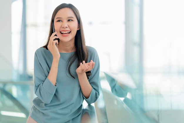 Lächelnde asiatische weibliche jugendlich Hand unter Verwendung des Smartphone-Gesprächs mit engem Freund im Kaufhaus des Einkaufszentrums oder im College-Korridor fröhliches Lachen, Lächeln, positive Einstellung, Emotion, junge asiatische Frau, Smartphone
