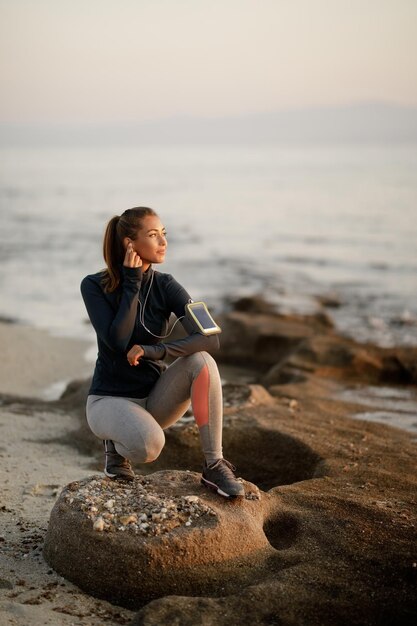 Lächelnde athletische Frau, die Musik und Tagträume auf einem Strandfelsen genießt Kopieren Sie Platz