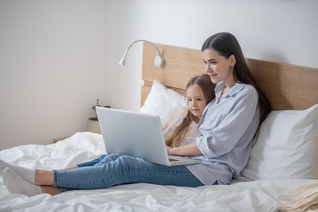 Lächelnde attraktive Frau und ihr süßes ruhiges Kind sitzen auf dem Bett vor dem Notebook-Computer