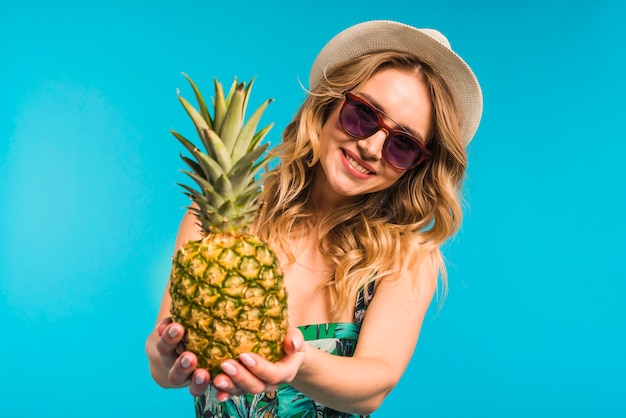 Lächelnde attraktive junge Frau im Hut und in Sonnenbrille, die frische Ananas halten