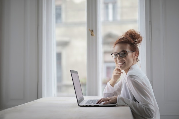 Lächelnde attraktive junge Frau mit Brille, die vor dem Laptop unter den Lichtern sitzt