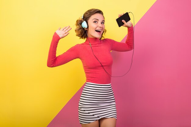 Lächelnde attraktive lächelnde aufgeregte Frau im stilvollen bunten Outfit tanzen und Musik in Kopfhörern auf rosa gelbem Hintergrund, Modesommertrend hören