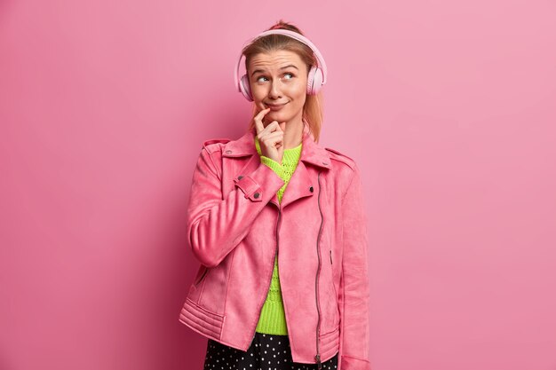 Lächelnde attraktive tausendjährige Frau mit verträumtem Ausdruck hört Lieblingslied, trägt Kopfhörer, genießt Wiedergabeliste, trägt rosa Jacke, steht drinnen. Hobby, Freizeit, Lifestyle