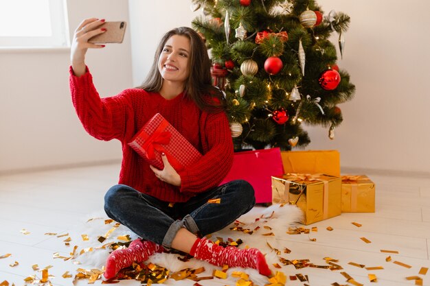 Lächelnde aufgeregte hübsche Frau im roten Pullover, der zu Hause am Weihnachtsbaum sitzt, der Geschenke und Geschenkboxen auspackt, die Selfie-Foto auf Telefonkamera nehmen