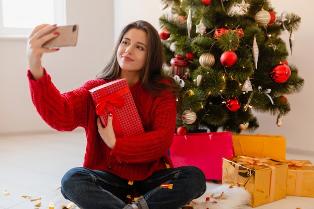 Lächelnde aufgeregte hübsche Frau im roten Pullover, der zu Hause am Weihnachtsbaum sitzt, der Geschenke und Geschenkboxen auspackt, die Selfie-Foto auf Telefonkamera nehmen