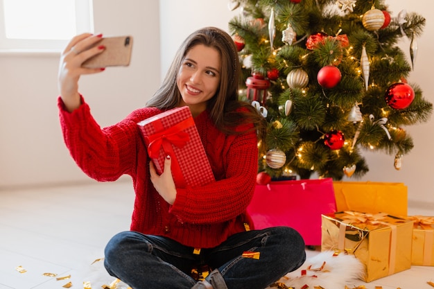 Lächelnde aufgeregte hübsche Frau im roten Pullover, der zu Hause am Weihnachtsbaum sitzt, der Geschenke und Geschenkboxen auspackt, die Selfie-Foto auf Telefonkamera nehmen