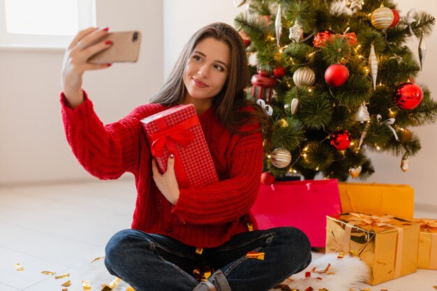 Lächelnde aufgeregte hübsche Frau im roten Pullover, der zu Hause am Weihnachtsbaum sitzt, der Geschenke und Geschenkboxen auspackt, die Selfie-Foto auf Telefonkamera nehmen