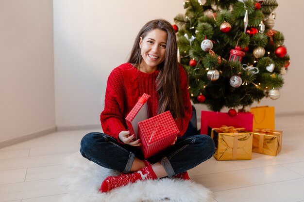 Lächelnde aufgeregte hübsche Frau im roten Pullover, der zu Hause am Weihnachtsbaum sitzt, der Geschenke und Geschenkboxen auspackt