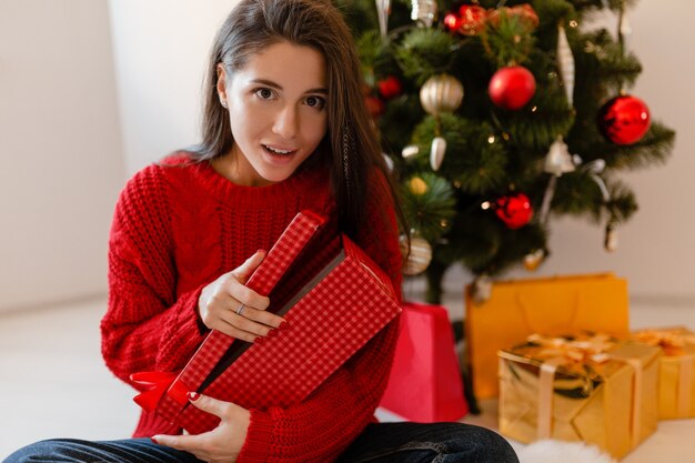 Lächelnde aufgeregte hübsche Frau im roten Pullover, der zu Hause am Weihnachtsbaum sitzt, der Geschenke und Geschenkboxen auspackt