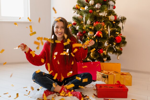 Lächelnde aufgeregte hübsche Frau im roten Pullover, der zu Hause am Weihnachtsbaum sitzt und goldenes Konfetti wirft, umgeben von Geschenken und Geschenkboxen