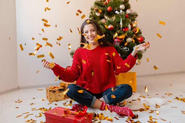 Lächelnde aufgeregte hübsche Frau im roten Pullover, der zu Hause am Weihnachtsbaum sitzt und goldenes Konfetti wirft, umgeben von Geschenken und Geschenkboxen