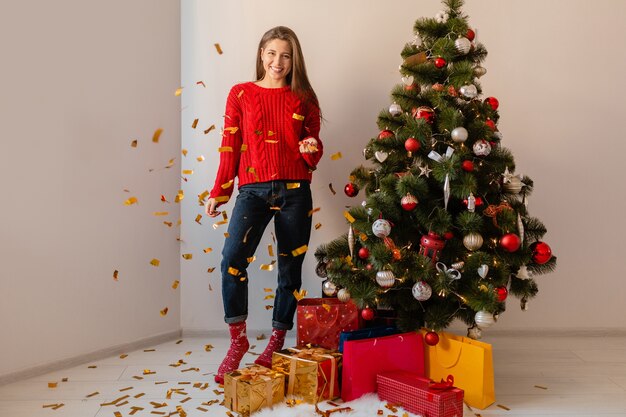 Lächelnde aufgeregte hübsche Frau im roten Pullover, der zu Hause am Weihnachtsbaum sitzt und goldenes Konfetti wirft, umgeben von Geschenken und Geschenkboxen