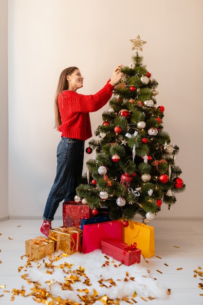 Lächelnde aufgeregte hübsche Frau im roten Pullover, der zu Hause steht und Weihnachtsbaum verziert, der mit Geschenken und Geschenkboxen umgeben ist