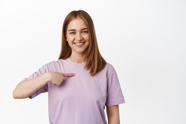 Lächelnde blonde Frau, die auf sich selbst zeigt, sich freiwillig erbietet, zur Teilnahme auffordert, prahlt, über ihre Leistungen spricht, über weiße Wand steht.