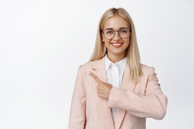Lächelnde blonde Frau in Gläsern, die mit dem Finger nach links zeigen, eine Brille und einen rosa Anzug auf Weiß tragen.