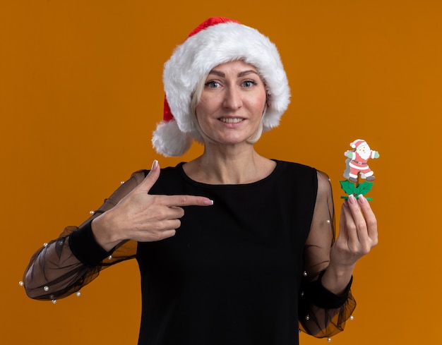 Lächelnde blonde Frau mittleren Alters mit Weihnachtsmütze, die das Weihnachtsmann-Spielzeug isoliert auf orangefarbener Wand hält und zeigt