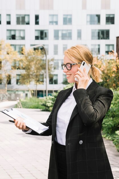 Lächelnde blonde junge Frau, die am Handy betrachtet Diagrammdiagramm auf Klemmbrett spricht