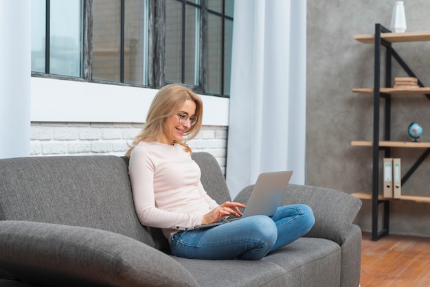 Lächelnde blonde junge Frau, die auf dem Sofa zu Hause schreibt auf Laptop sitzt