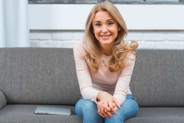 Lächelnde blonde junge frau, die auf grauem sofa mit laptop sitzt