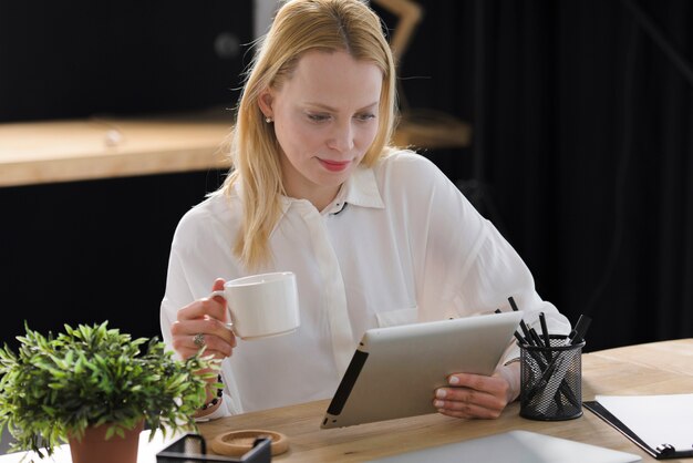 Lächelnde blonde junge Frau, die Tasse Kaffee betrachtet digitale Tablette hält