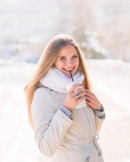 Lächelnde blonde junge Frau, die wegwerfbare Kaffeetasse im Winter trinkt