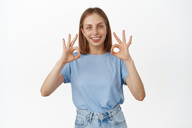 Lächelnde blonde Kaukasierin zeigt okay OK-Zeichen, genehmigt etwas Cooles, lobt großartige Arbeit, empfiehlt Produkt, steht über weißer Wand